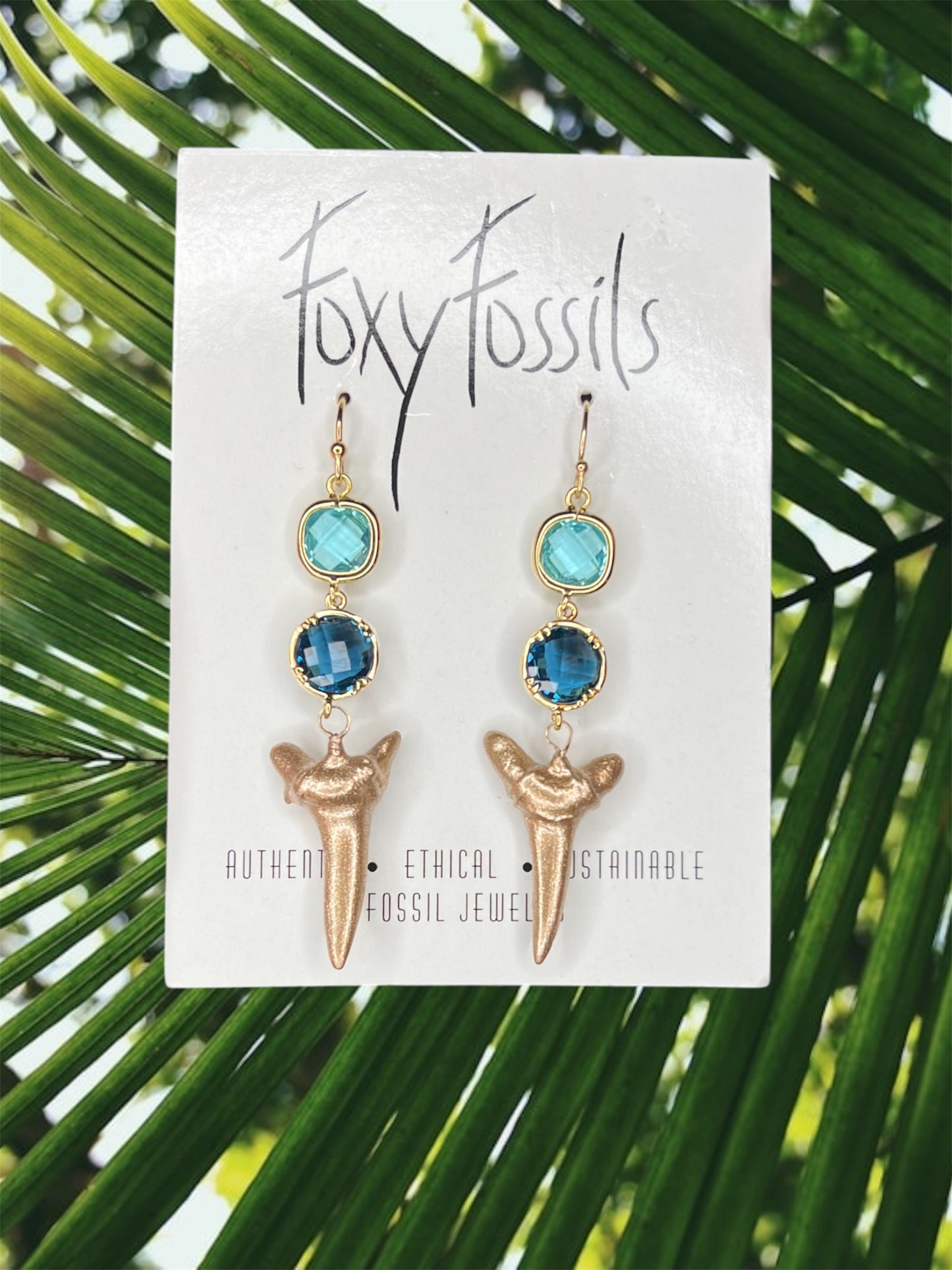 A pair of gold-painted real fossil shark teeth handmade dangle earrings with blue framed glass connectors, made by Foxy Fossils using ethically and legally collected fossils.