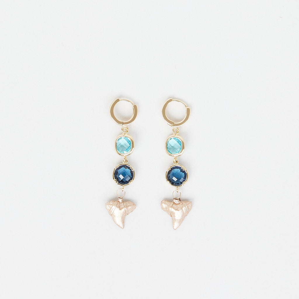 Pair of earrings with gold hoops, blue gemstones, and real gold fossil shark teeth on a white background.